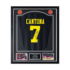 Vooraanzicht van een zwart Manchester United 1994-1996 uitshirt, gesigneerd door Eric Cantona op geel rugnummer 7, ingelijst in een zwart-zilveren Deluxe Frame met twee actiefoto's.